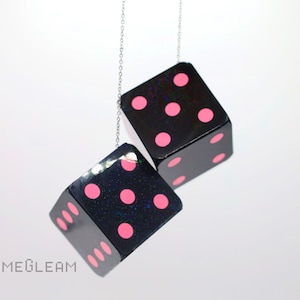 May include: Two black dice with pink dots hanging from a silver chain. The dice are hanging against a white background.