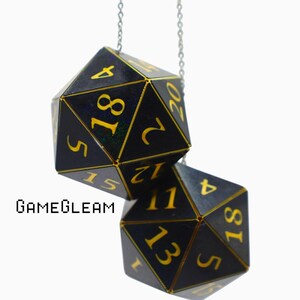 May include: Two black twenty-sided dice pendants hanging from a silver chain. Each die features gold-colored numbers and edges. The text GAMEGLEAM is printed in black to the left of the lower die. The dice have a smooth, dark finish with gold numerals.