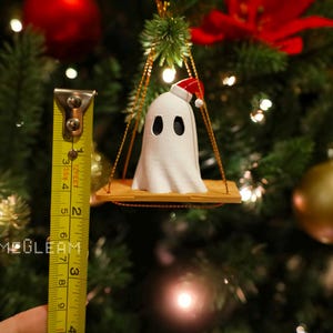 A close up view of a white Santa ghost on a brown swing ornament hanging on a holiday Christmas tree with lights in the background. A yellow measuring tape shows the ornament is 2 inches tall.