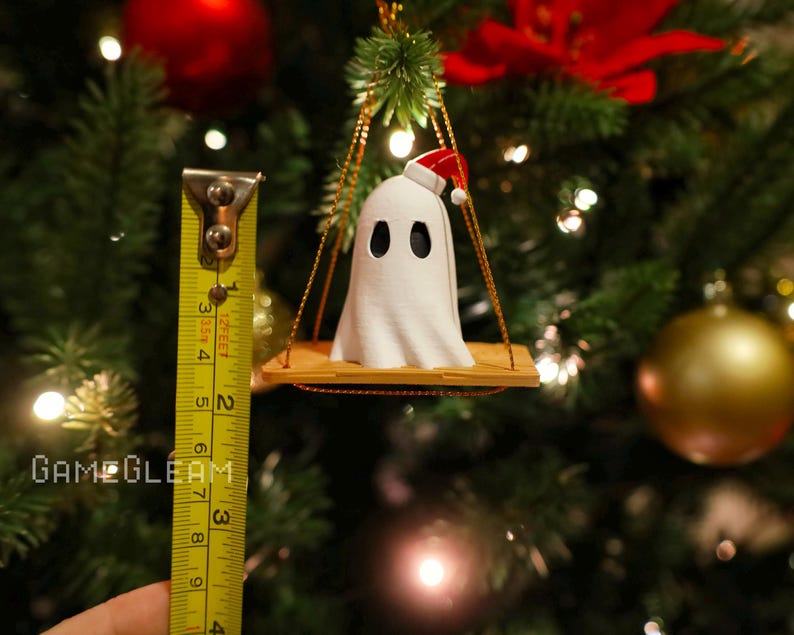 A close up view of a white Santa ghost on a brown swing ornament hanging on a holiday Christmas tree with lights in the background. A yellow measuring tape shows the ornament is 2 inches tall.