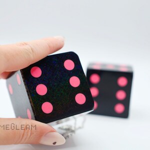 A close up view of two holographic black car dice with pink dot pips with a linked silver chain. The die is being held by a model which shows the die fits in her palm.