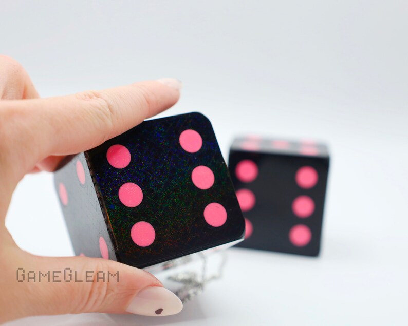 A close up view of two holographic black car dice with pink dot pips with a linked silver chain. The die is being held by a model which shows the die fits in her palm.