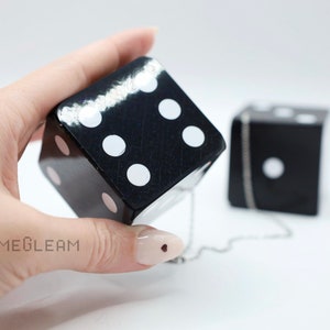 A close up view of two holographic black car dice with white dot pips with a linked silver chain. The die is being held by a model which shows the die fits in her palm.
