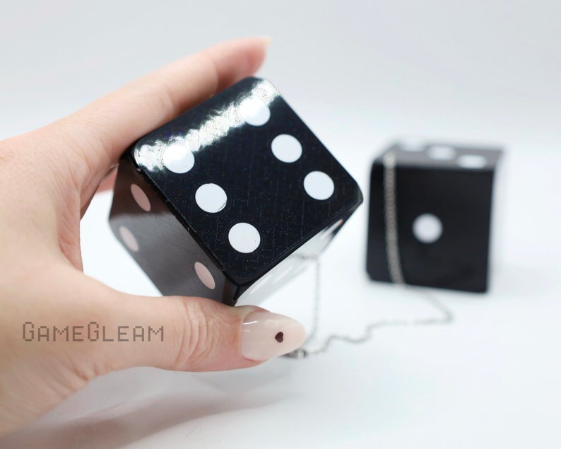 A close up view of two holographic black car dice with white dot pips with a linked silver chain. The die is being held by a model which shows the die fits in her palm.