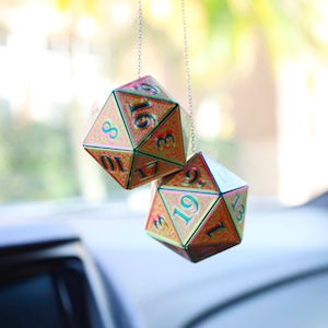 May include: Two iridescent dice hanging from a silver chain. The dice are gold and pink with black numbers. The dice are a 20-sided die and a 12-sided die.