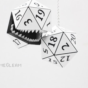 May include: Two white twenty-sided dice with black numbers and black outlines. One die is open, revealing a set of sharp white teeth inside the hollow structure. The text GAMEGLEAM is printed in black in the lower left corner of the image.