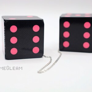 A close up side view of two holographic black car dice with pink dot pips and a linked silver chain laying on a white surface.