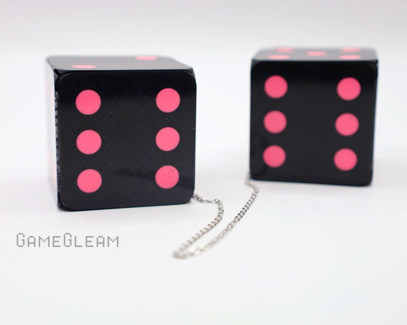 A close up side view of two holographic black car dice with pink dot pips and a linked silver chain laying on a white surface.