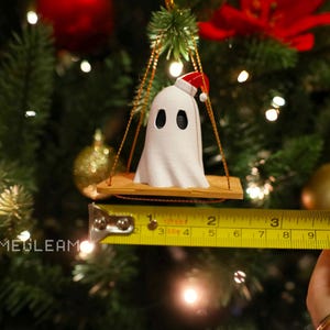 A close up view of a white Santa ghost on a brown swing ornament hanging on a holiday Christmas tree with lights in the background. A yellow measuring tape shows the ornament is 2 inches wide.