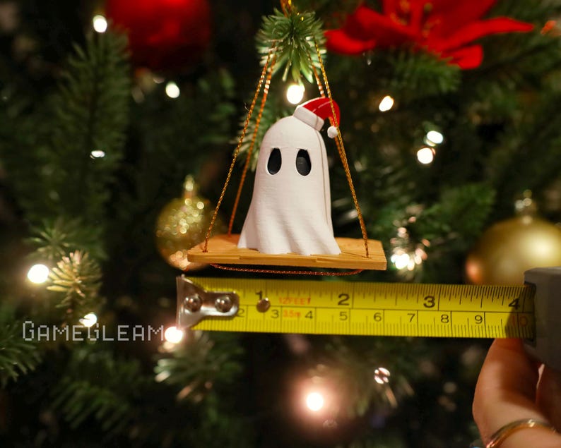 A close up view of a white Santa ghost on a brown swing ornament hanging on a holiday Christmas tree with lights in the background. A yellow measuring tape shows the ornament is 2 inches wide.