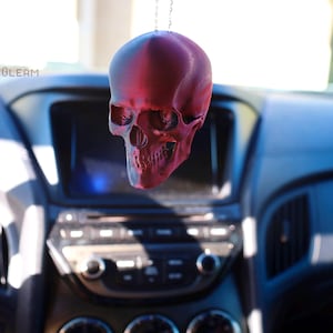 May include: A red plastic skull hanging from a rearview mirror in a car. The skull is detailed with eye sockets, a nose, and teeth.