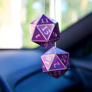 May include: Two purple and gold dice with numbers 1, 5, 11, 13, 18, 20 hanging from a silver chain. The dice are hanging from a rearview mirror in a car.