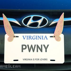 May include: A white Virginia license plate with the text "PWNY" and the slogan "Virginia is for lovers". Two pink and gold butterfly-shaped ornaments are attached to the plate.