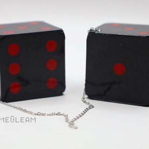 A close up side view of two holographic black car dice with red dot pips and a linked silver chain laying on a white surface.
