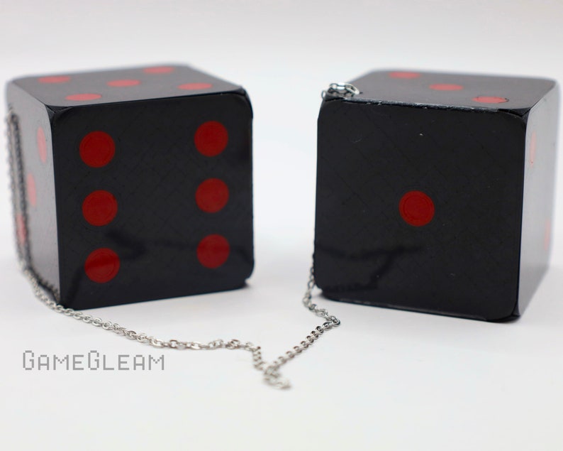 A close up side view of two holographic black car dice with red dot pips and a linked silver chain laying on a white surface.