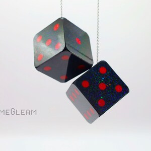 A close up side view of two holographic black car dice with red dot pips and a linked silver chain hanging over a white surface.