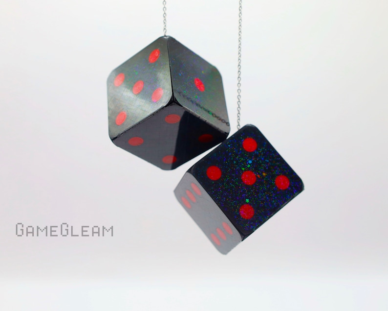 A close up side view of two holographic black car dice with red dot pips and a linked silver chain hanging over a white surface.