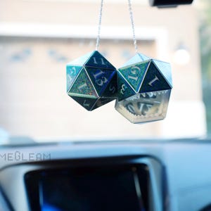 May include: Two iridescent blue and green dice hanging from a rearview mirror in a car. The dice are shaped like 20-sided polyhedrons and have white numbers on each side.