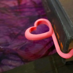 May include: A pink heart-shaped bookmark made of plastic. The bookmark is holding a book closed.