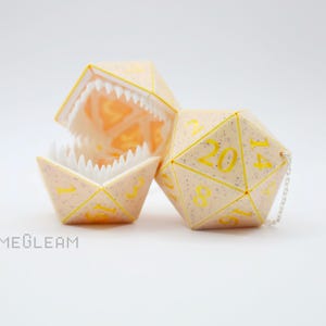May include: A set of two white dice with yellow numbers. One die is a d20 with a chain attached. The other die is a d20 with a hinged jaw that opens to reveal the inside.
