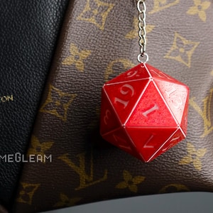 Pode incluir: Um pingente de dados vermelho de vários lados com numeração branca, pendurado em uma corrente prateada. O dado é um acessório de jogo. O fundo inclui uma bolsa estampada marrom e preta.