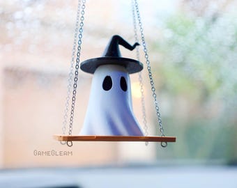 Op de afbeelding: Een witte geest met een zwarte heksenhoed die aan een zilveren ketting hangt. De geest zit op een kleine houten schommel.