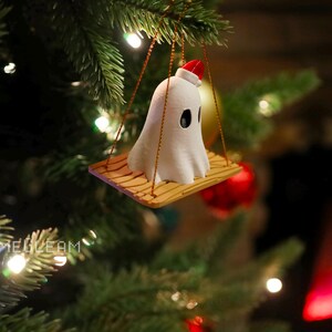 A top down view of a white Santa ghost on a brown swing ornament hanging on a holiday Christmas tree with lights in the background.