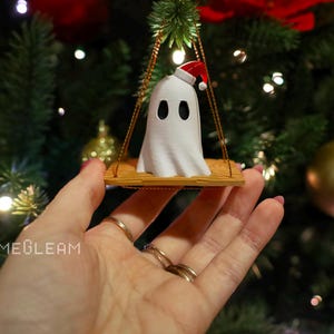 A close up view of a white Santa ghost on a brown swing ornament hanging on a holiday Christmas tree with lights in the background. A model has her hand holding the ornament.
