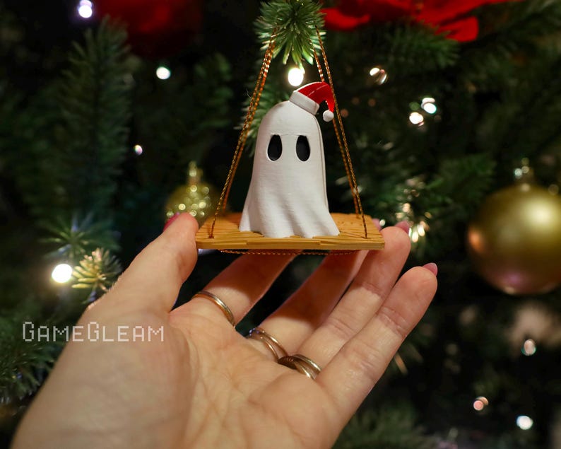 A close up view of a white Santa ghost on a brown swing ornament hanging on a holiday Christmas tree with lights in the background. A model has her hand holding the ornament.