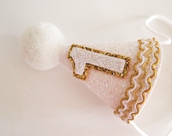 May include: White felt party hat with a gold glitter trim and a white felt number one with gold glitter outline. The hat has a white pom-pom on top and a white string.