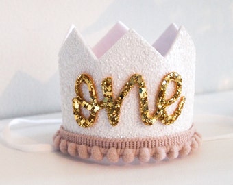 May include: White glitter crown with a gold glitter "one" on the front. The crown has a pink pom-pom trim.
