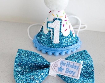 May include: A blue glitter bow tie with a white tag that says "Birthday Boy" and a blue glitter party hat with a white pom pom and the number "1" on it.