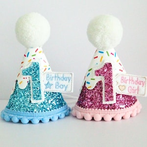 May include: Two party hats with white pom poms on top. The hats are decorated with colourful sprinkles and glitter. One hat is blue with a white number one and the text "Birthday Boy". The other hat is pink with a white number one and the text "Birthday Girl".