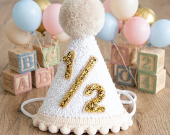 May include: A white textured cone-shaped party hat featuring a large gold glitter fraction 1/2 on the front, topped with a beige pom-pom and trimmed with small beige pom-poms at the base, accompanied by pastel balloons and wooden alphabet blocks.