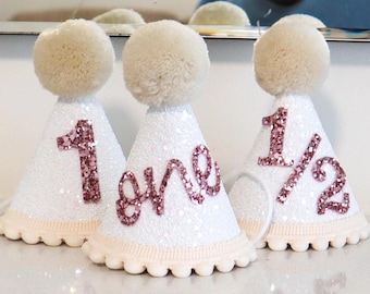 May include: Three white party hats with pink glitter accents. The hats are decorated with the numbers "1", "one", and "1/2".
