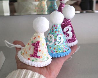 May include: Three small cone-shaped party hats featuring glittery surfaces and white pom-poms on top. The first hat is white with a pink glittery number 1, the second is light blue with white number 99, and the third is purple with a white number 3.