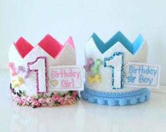 May include: Two handmade birthday crowns, one pink and one blue, with the number "1" and the words "Birthday Girl" and "Birthday Boy". The crowns are white with glitter, and decorated with flowers and bows.