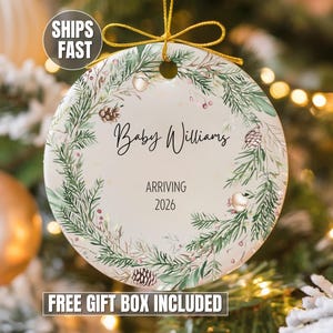 May include: A round ceramic Christmas ornament with a festive wreath design. The ornament features the text "Baby Williams ARRIVING 2026". The wreath includes green foliage, berries, and pinecones. A gold ribbon is attached for hanging.