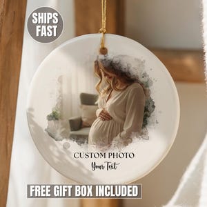May include: A round white ceramic ornament featuring a watercolor-style printed photograph of a pregnant person in a light-colored dress. The ornament includes a gold hanging string and the printed text CUSTOM PHOTO and Your Text in black ink.