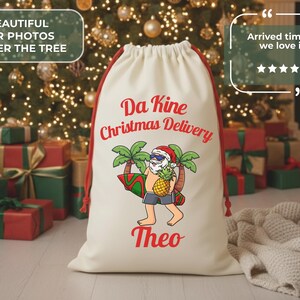 May include: Cream-colored Christmas gift bag with red drawstring and a Santa Claus graphic. The Santa is wearing sunglasses and holding a surfboard and pineapple. Text on the bag reads "Da Kine Christmas Delivery" and "Theo".