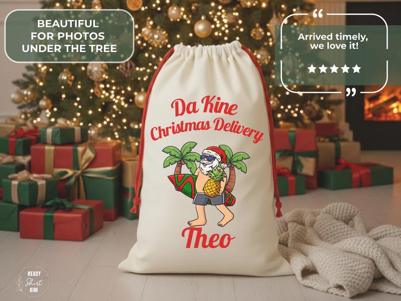 May include: Cream-colored Christmas gift bag with red drawstring and a Santa Claus graphic. The Santa is wearing sunglasses and holding a surfboard and pineapple. Text on the bag reads "Da Kine Christmas Delivery" and "Theo".