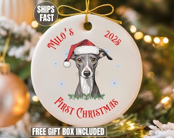 May include: A round white ceramic ornament featuring a grey and white Italian Greyhound wearing a red Santa hat. The text reads Milo's 2025 First Christmas in red lettering. The ornament includes a gold string for hanging and green holly accents.