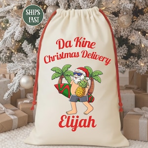 May include: Cream-colored drawstring gift bag with red accents. The bag features a cartoon Santa Claus with sunglasses, a pineapple, and a surfboard. Text on the bag reads "Da Kine Christmas Delivery Elijah."