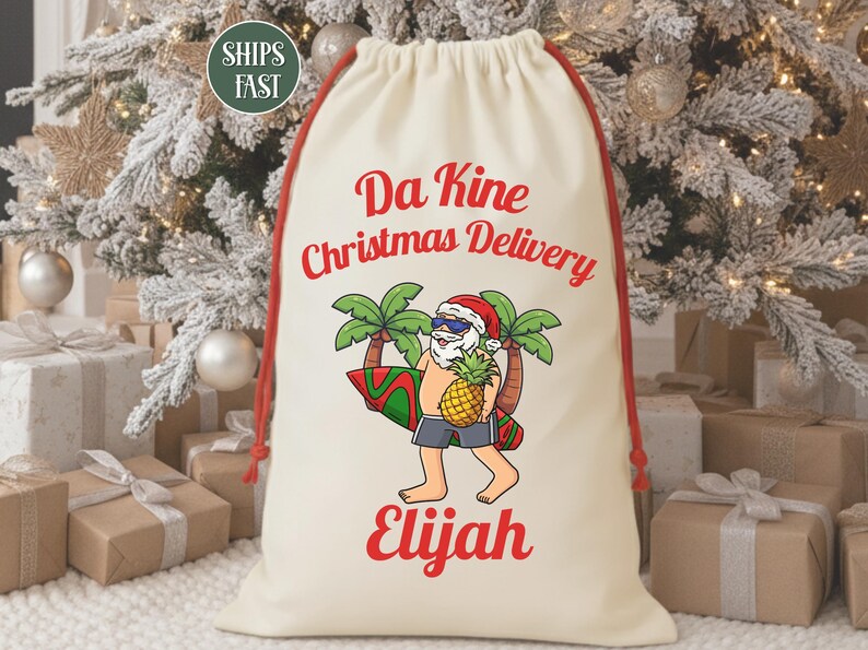 May include: Cream-colored drawstring gift bag with red accents. The bag features a cartoon Santa Claus with sunglasses, a pineapple, and a surfboard. Text on the bag reads "Da Kine Christmas Delivery Elijah."