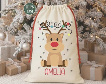 May include: A cream-colored Christmas gift bag with a cartoon reindeer design. The reindeer has a red nose and is wearing string lights. The name "AMELIA" is printed in red. The bag has a red drawstring.