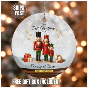 May include: A round Christmas ornament featuring a family of three nutcracker figures. The ornament has the text "Our First Christmas as a Family of Three 2025". The ornament is white with gold accents and a gold string.