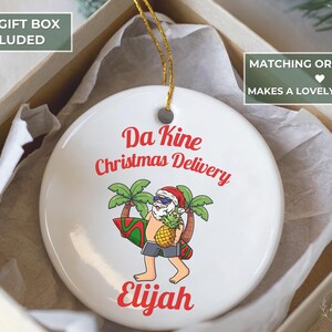 May include: White ceramic Christmas ornament with a cartoon Santa Claus carrying a surfboard and pineapple. The ornament has the text "Da Kine Christmas Delivery" and a name in red. It is packaged in a gift box.