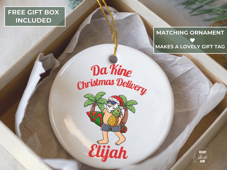 May include: White ceramic Christmas ornament with a cartoon Santa Claus carrying a surfboard and pineapple. The ornament has the text "Da Kine Christmas Delivery" and a name in red. It is packaged in a gift box.
