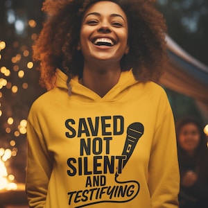 May include: A smiling person wearing a yellow hoodie with the text "SAVED NOT SILENT AND TESTIFYING" in black. The hoodie features a microphone graphic. The background is blurred with bokeh lights.