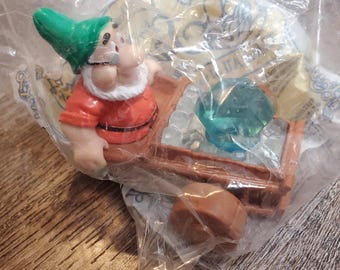 May include: A plastic toy figurine of a dwarf from Snow White and the Seven Dwarfs. The dwarf is wearing a red shirt, green hat, and white beard. He is pushing a wooden cart with a large green gem inside.